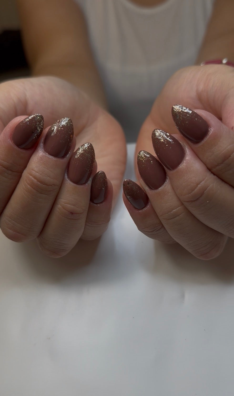 Close-up of hands with almond-shaped nails painted in a glossy taupe-brown shade with fine silver glitter tips.